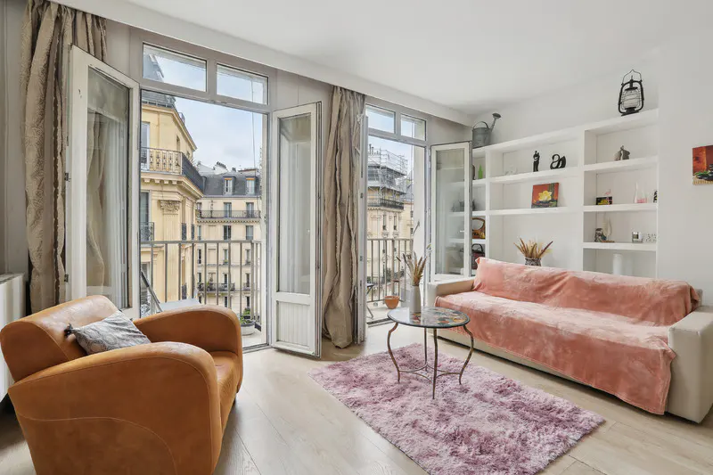 Bright living room with open balcony doors showing city view. A tan chair, pink rug, and white sofa with a pink throw blanket are visible.