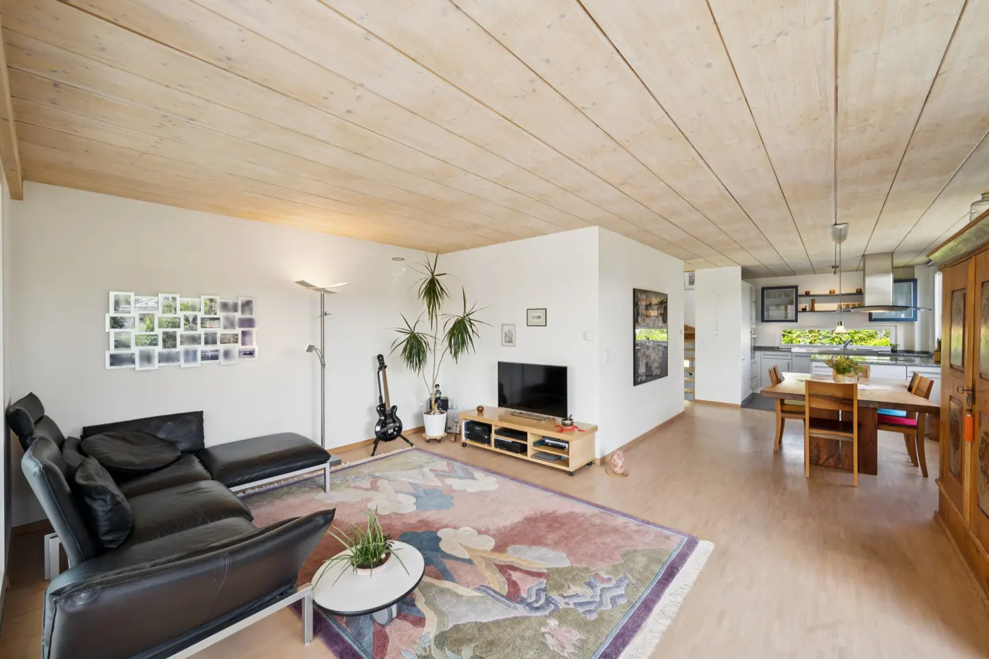 Open-concept living room with wood ceiling, black leather sofa, TV, and colorful rug. Kitchen and dining area visible in the background.