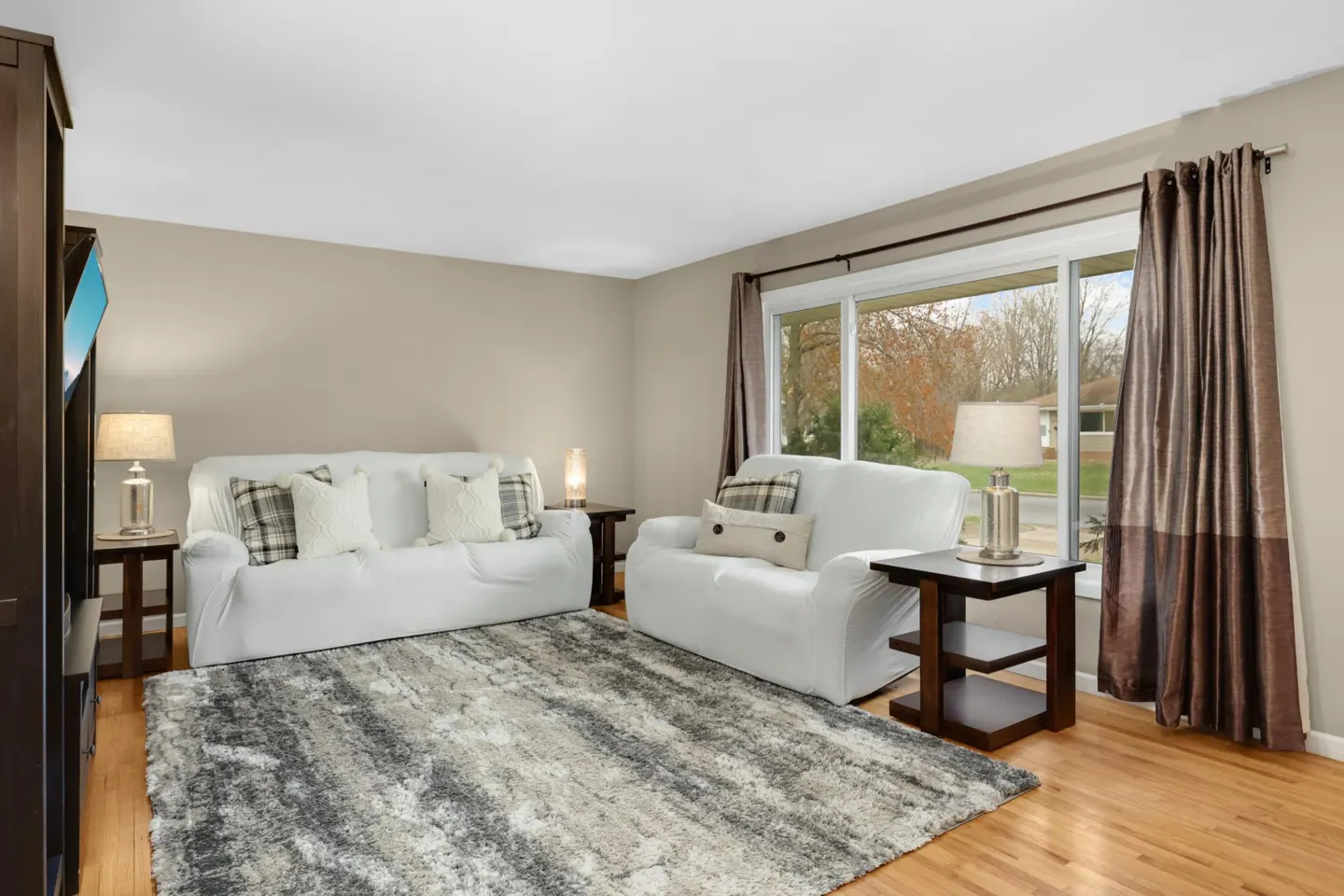 Living room with white sofas, gray rug, wood floors, and a large window with brown curtains.