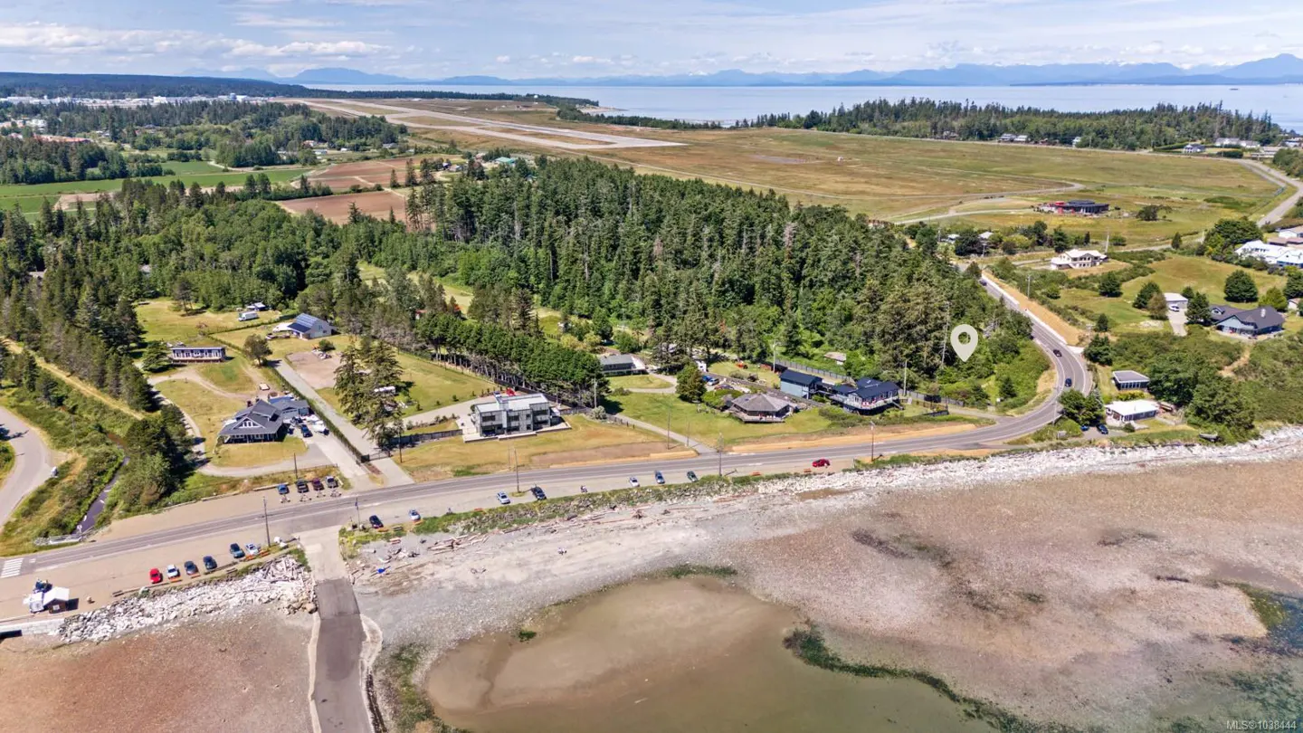 Aerial view of coastal real estate with homes, trees, a road, and a tidal beach. An airport runway is visible in the background.