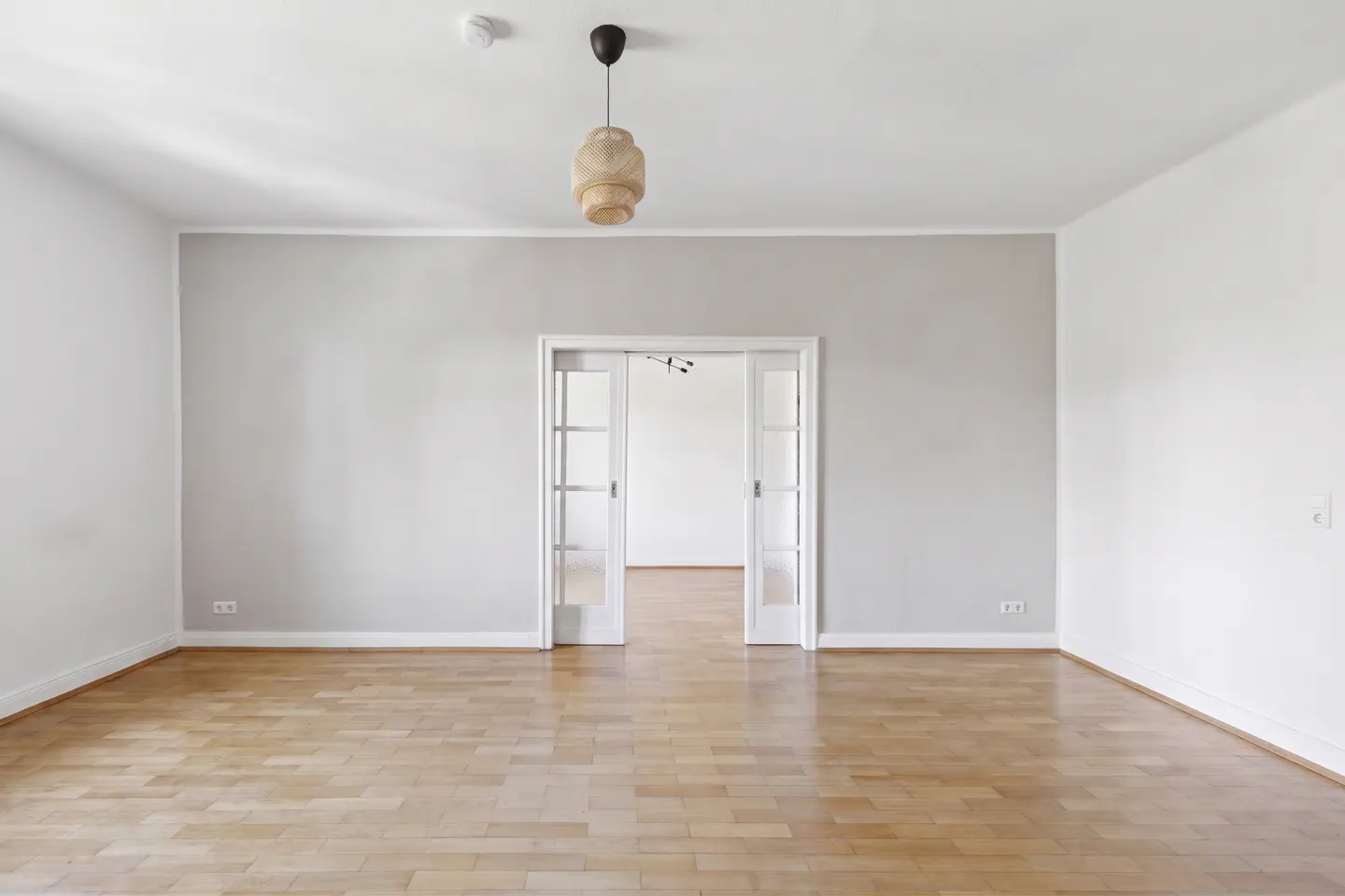 Empty room with light wood floors, gray walls, and white trim. A woven pendant light hangs from the white ceiling. Glass doors lead to another room.