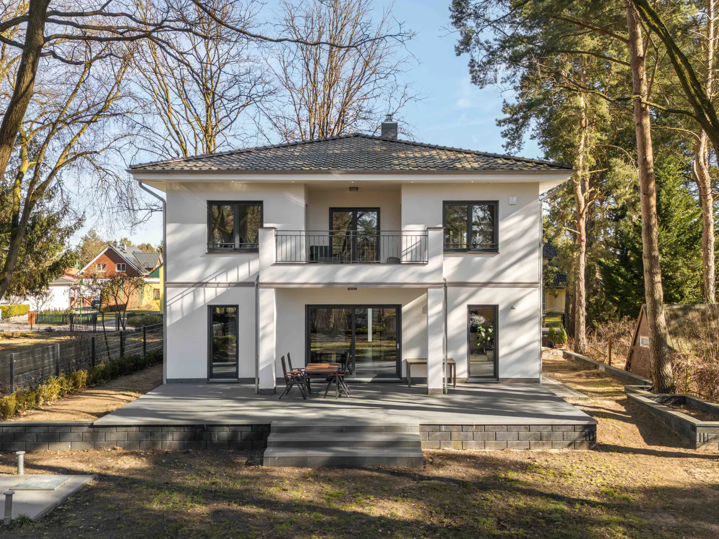 Two-story white house with a gray roof, black windows, and a balcony overlooking a gray patio with a table and chairs.
