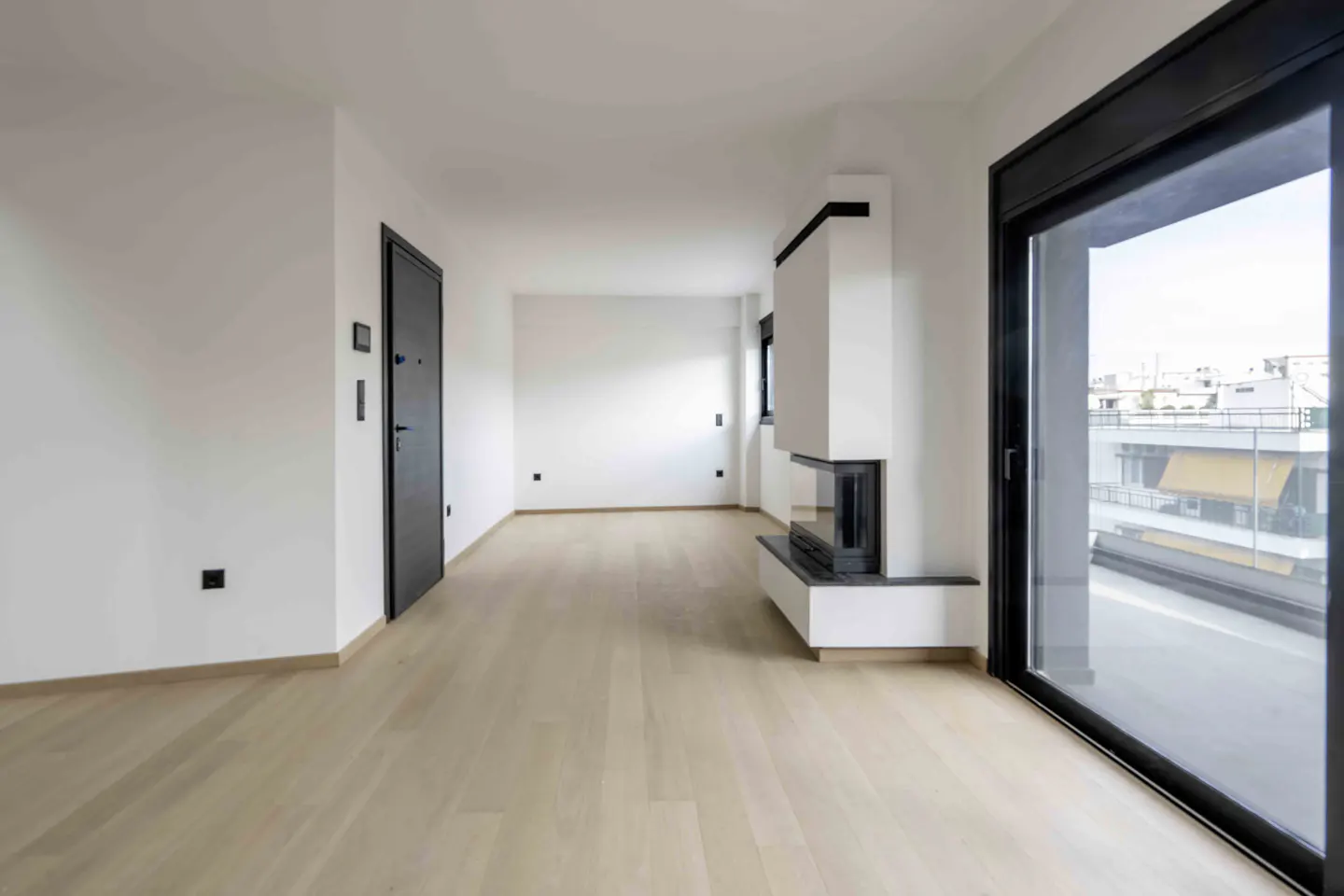 Bright, empty room with light wood floors, white walls, and a modern white fireplace. A large sliding glass door shows a city view.
