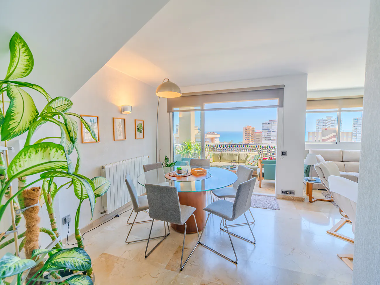 Bright dining area with a glass table, gray chairs, and a large window overlooking a balcony with ocean views. A large green plant is in the foreground.