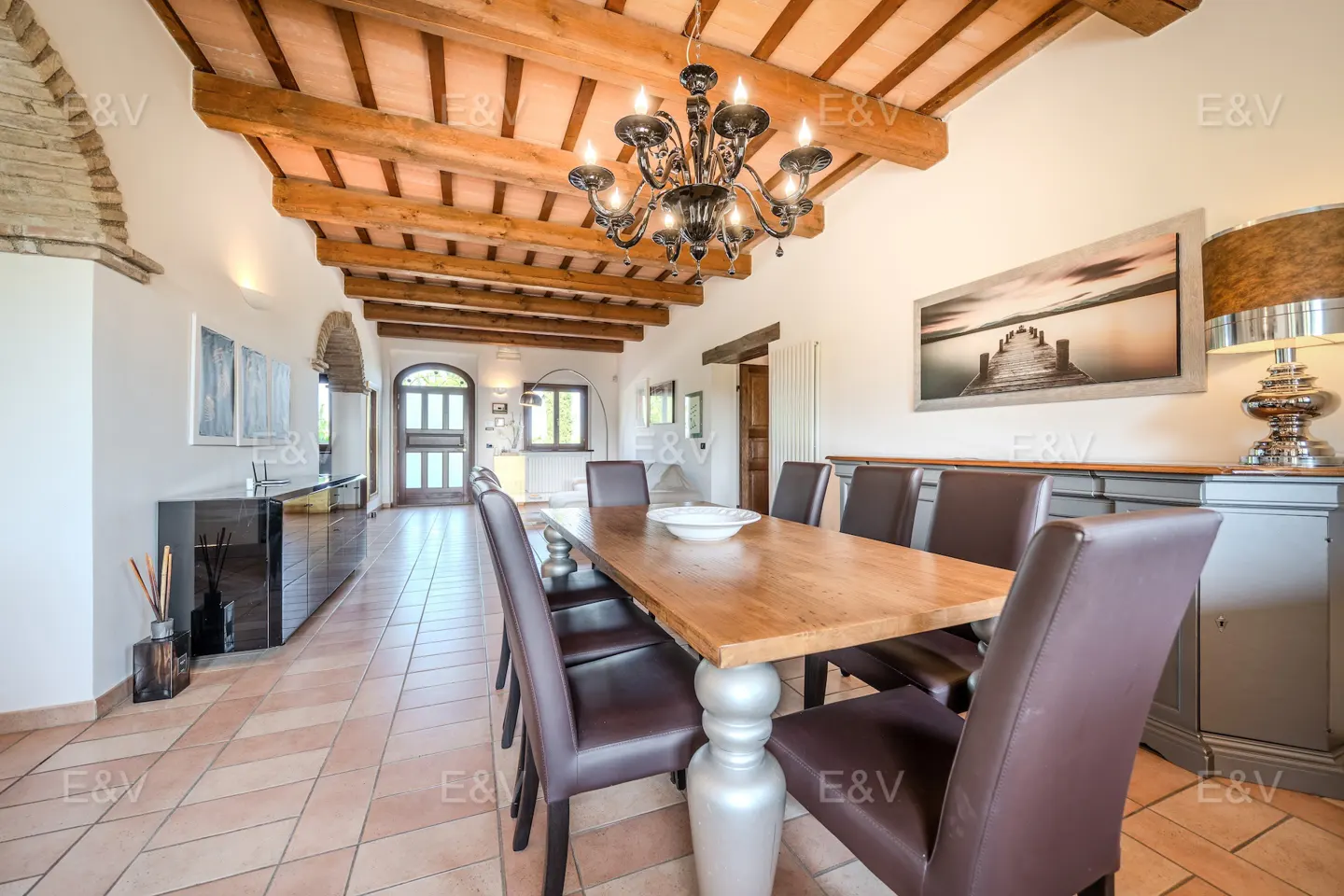 Dining room with a long wooden table, brown leather chairs, and a chandelier. The ceiling has exposed wooden beams. A landscape painting hangs on the wall.