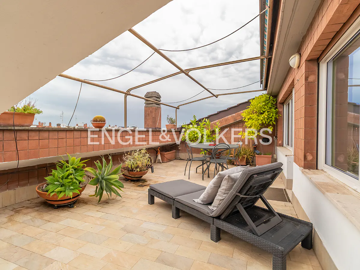 Spacious rooftop terrace with stone tiles, a lounge chair, dining set, potted plants, and a metal frame against a cloudy sky.