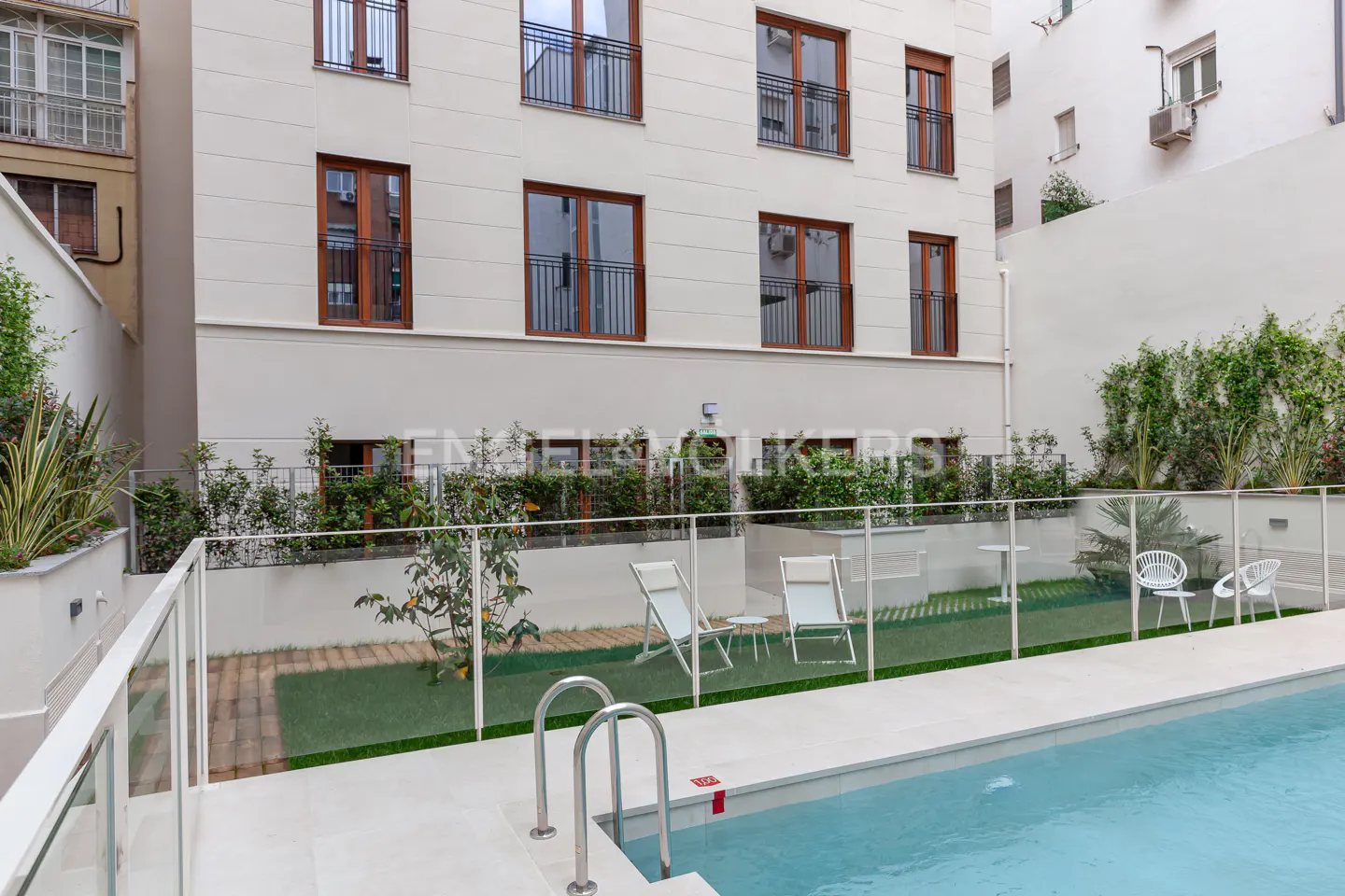 Apartment building with a pool. The pool has a metal ladder and is surrounded by a white deck and a glass fence. There are chairs and greenery.