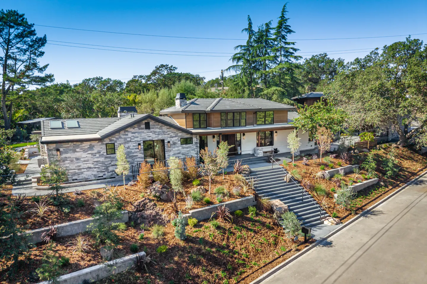 Two-story house with stone and wood facade, gray roof, and black-framed windows. Landscaped yard with tiered retaining walls and long staircase.