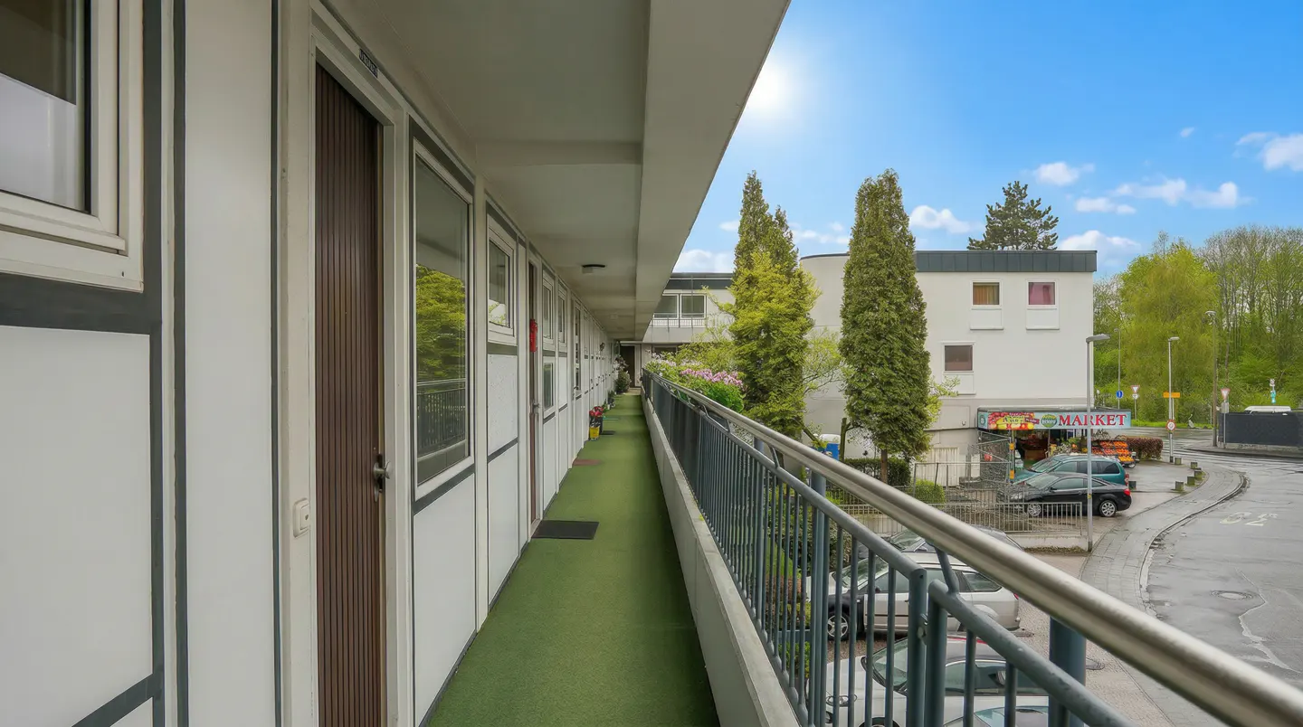 Apartment balcony with green carpet, metal railing, and view of a market across the street under a blue sky.