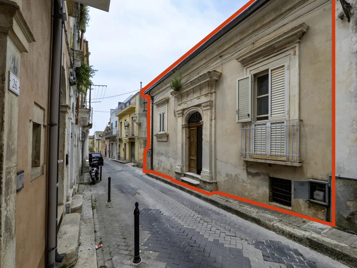 Exterior view of a beige building with a red outline, featuring a wooden door, a window with a balcony, and a narrow cobblestone street.