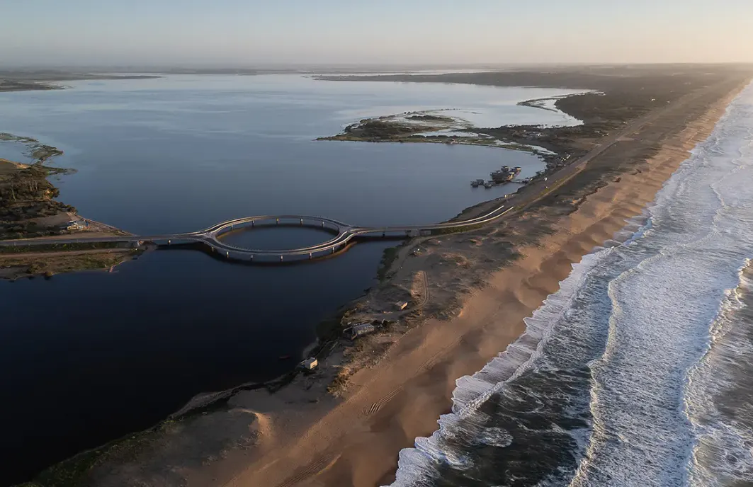 Aerial view of a sandy beach with waves, a lake, and a circular bridge connecting land masses.