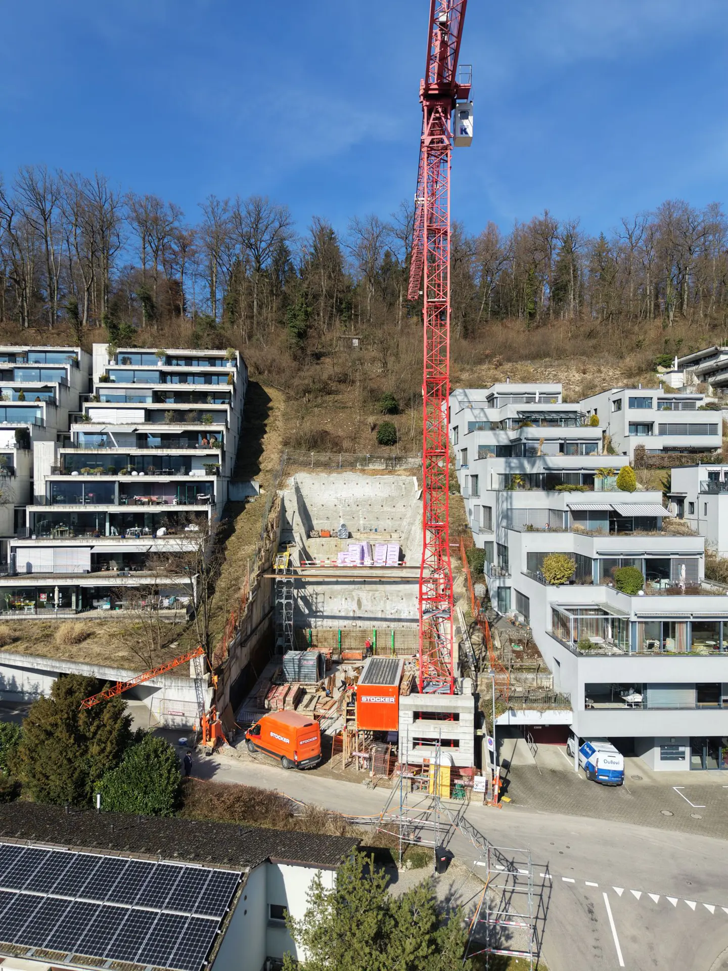 Construction site with a red crane, excavation, and modern terraced apartments on a hillside under a blue sky.