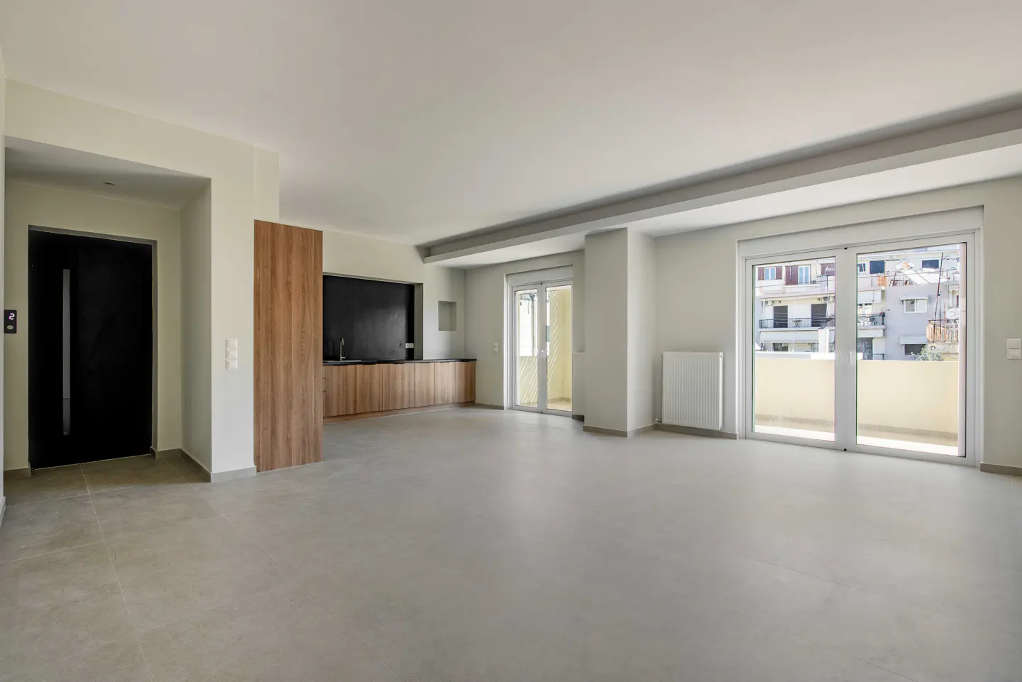 Bright, empty apartment with gray floors, white walls, and a black front door. A wood-paneled kitchen area is visible, with sliding glass doors leading to a balcony.