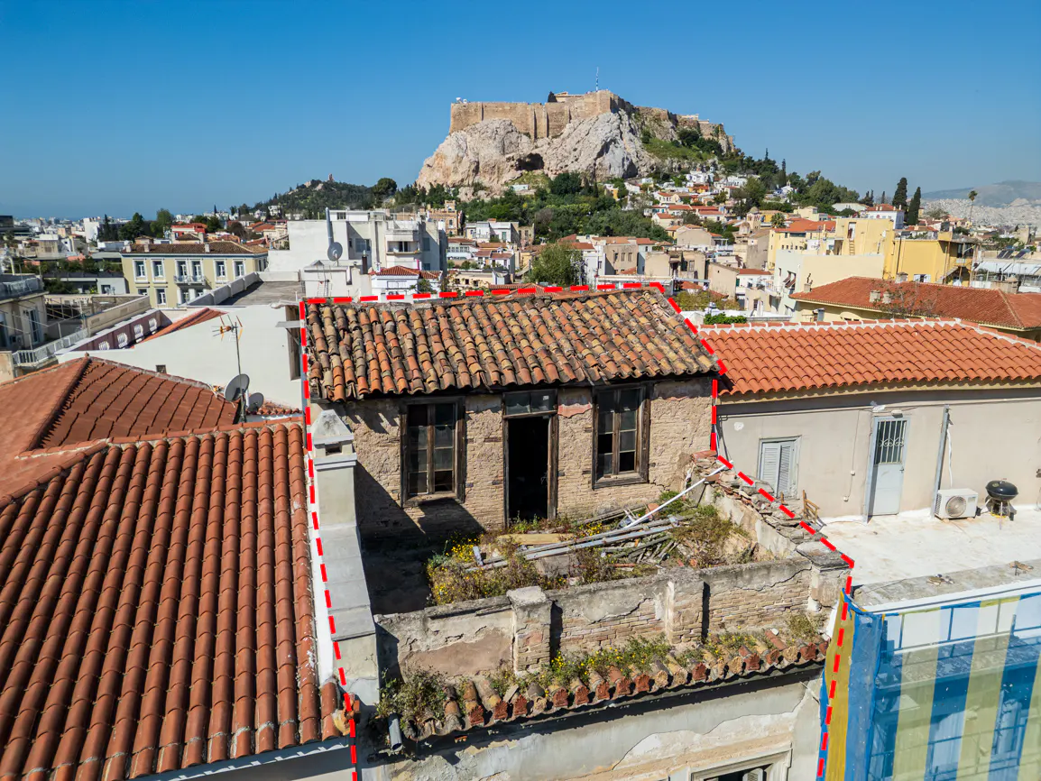 Athens rooftop view of a small, rundown building with a red-dashed outline, and the Acropolis in the background.