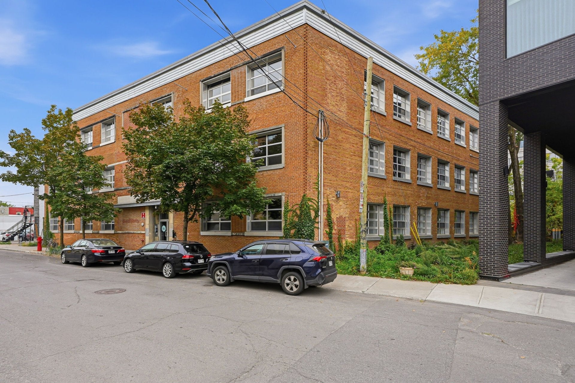 Three-story red brick building on a city street with parked cars, green trees, and a modern dark brick structure nearby. [AI-generated text]