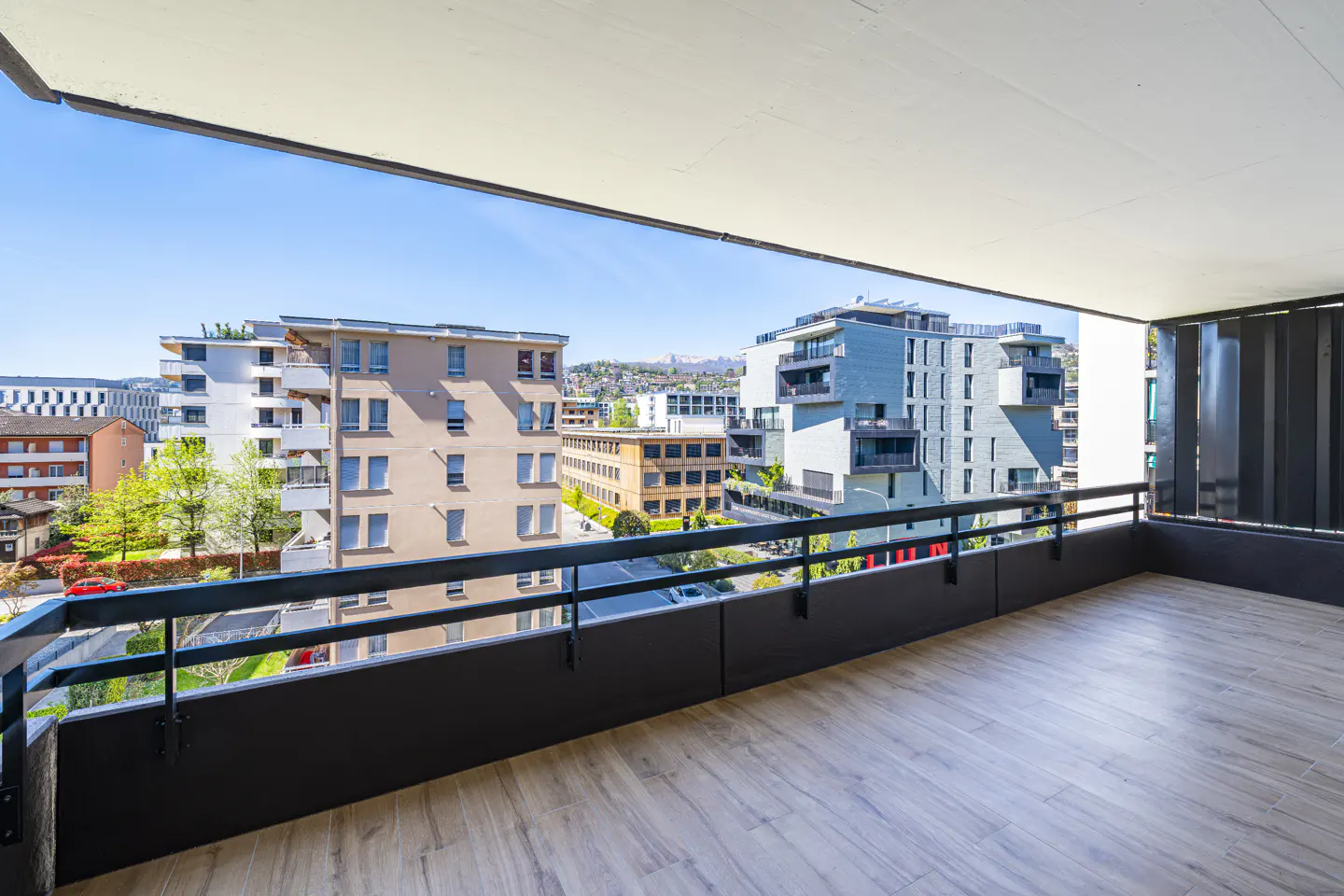 View from a balcony with a black railing, overlooking city buildings under a blue sky.
