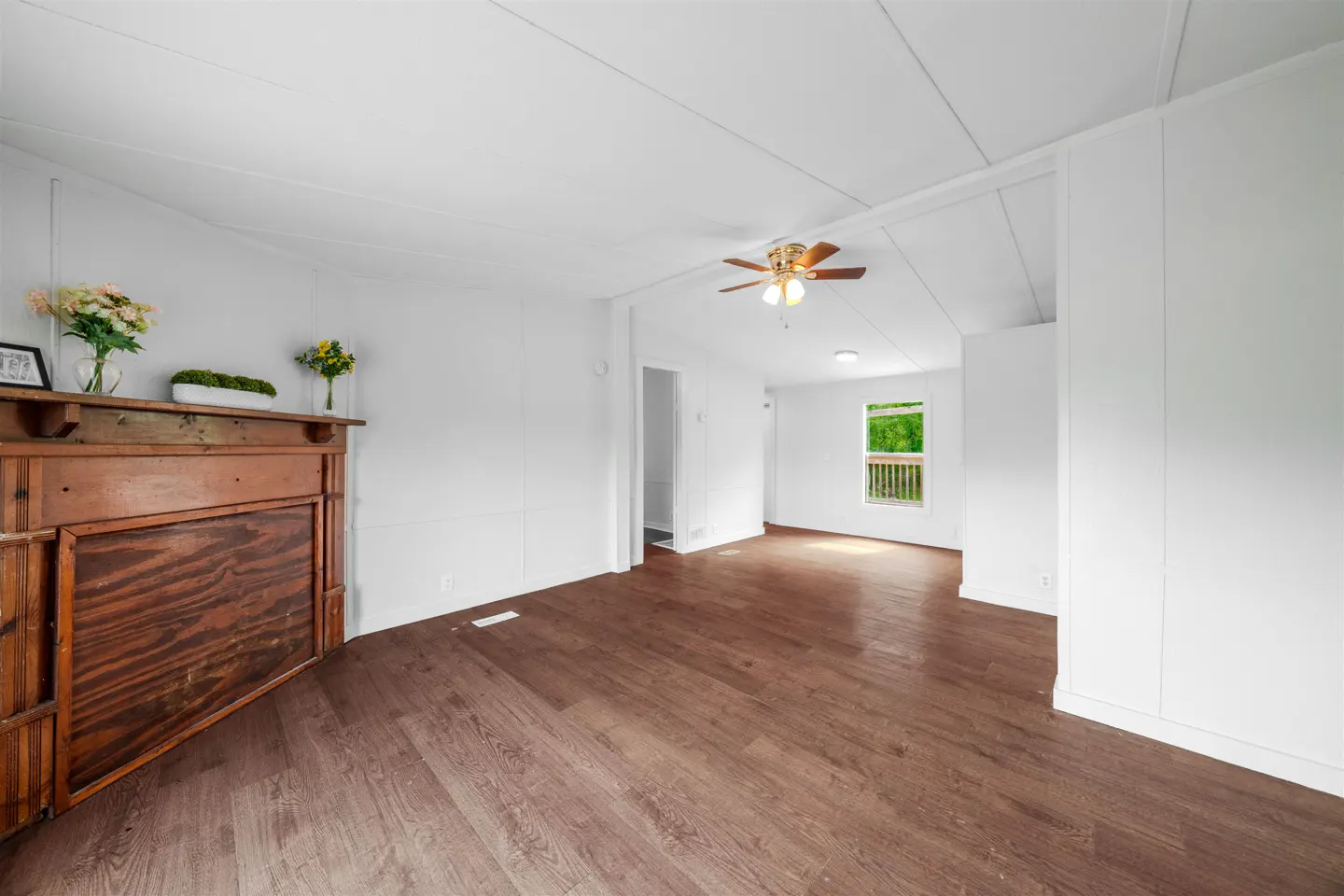 Bright living room with dark wood floors, white walls, and a brown fireplace with flowers on the mantle. A ceiling fan is visible.