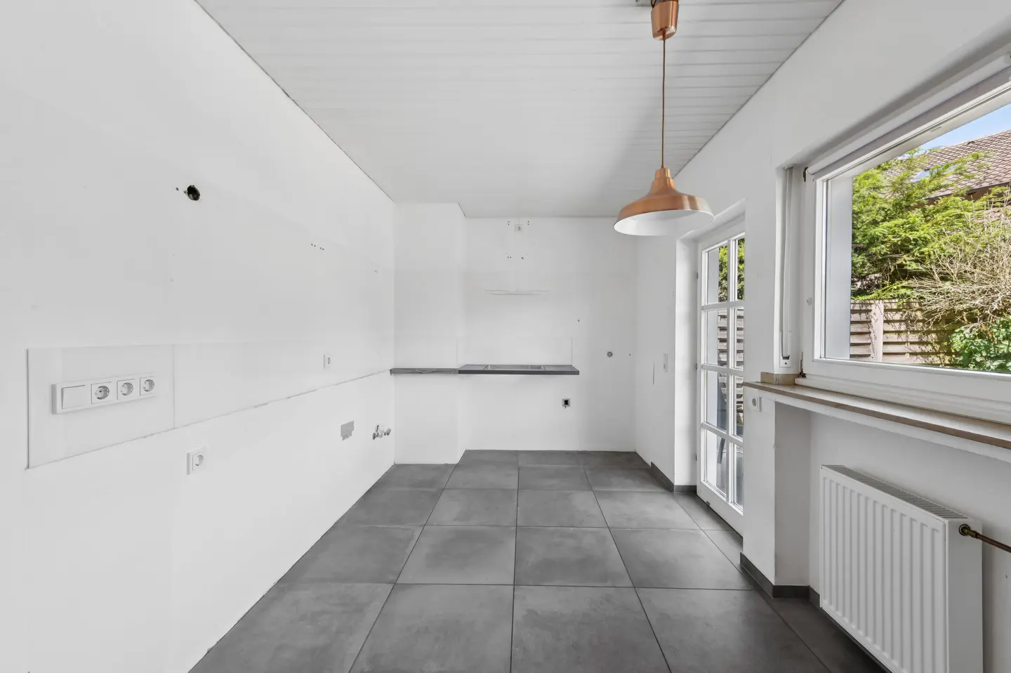 Empty kitchen with white walls, grey tiled floor, a copper pendant light, and a window looking out to a garden.