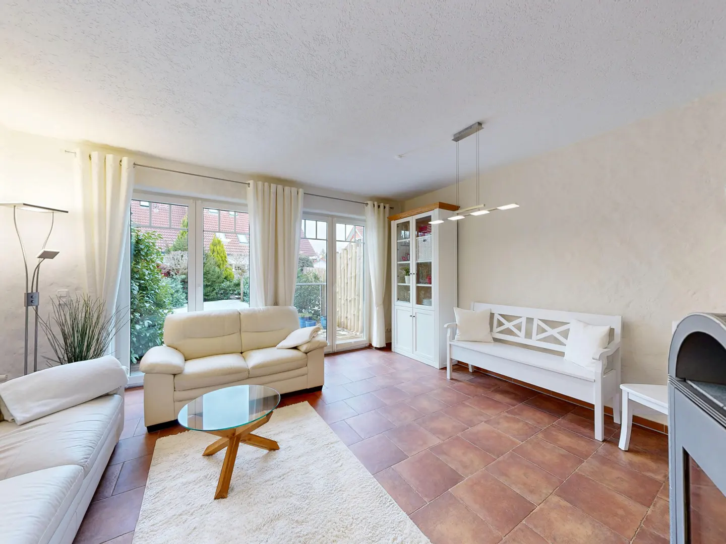 Living room with white sofas, glass table, and white bench. Brown tile floor and white rug. Large windows with garden view.