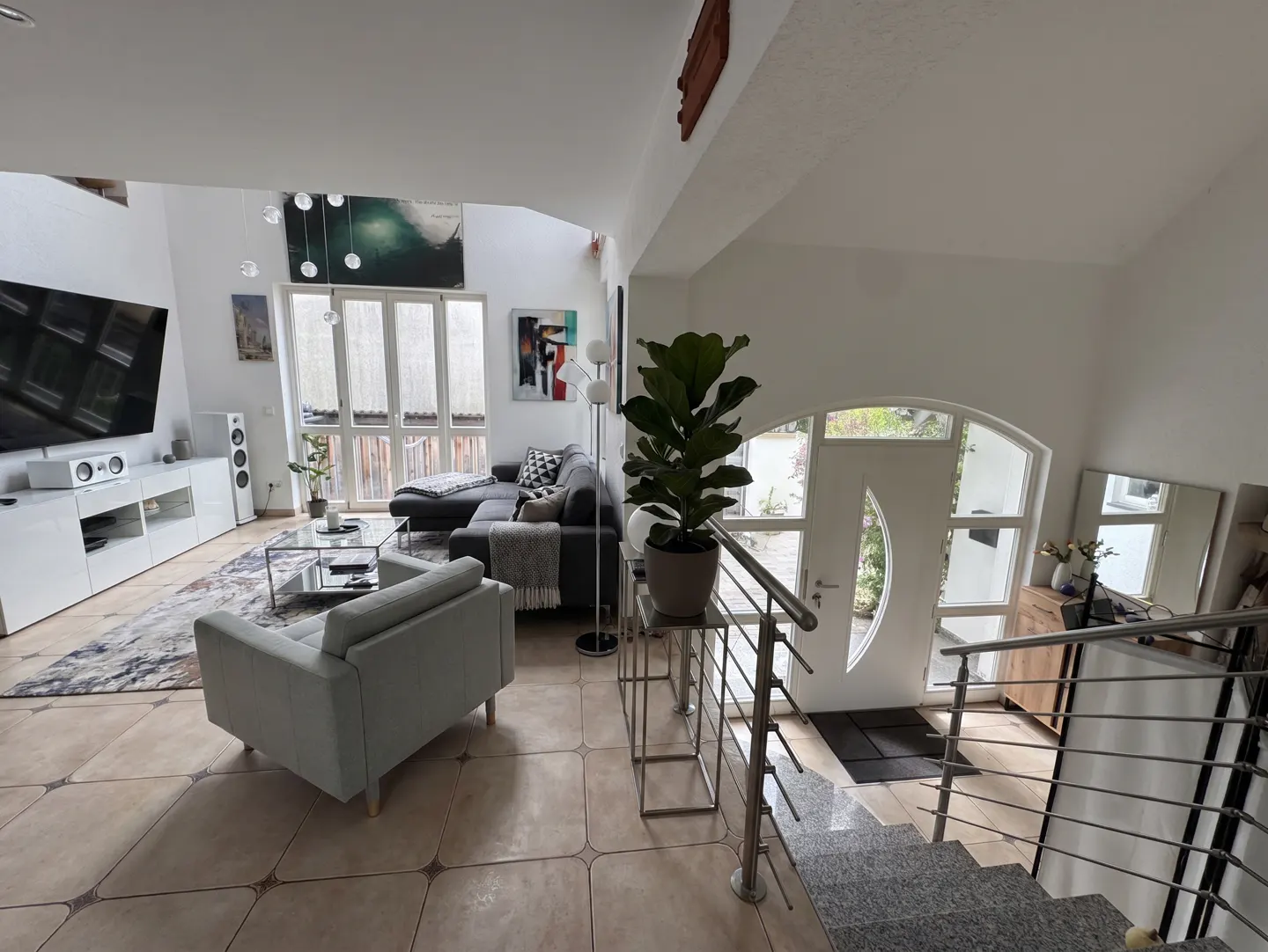 Bright, modern living room with a gray sofa, chair, and a large TV. A staircase with metal railings leads to a white front door.