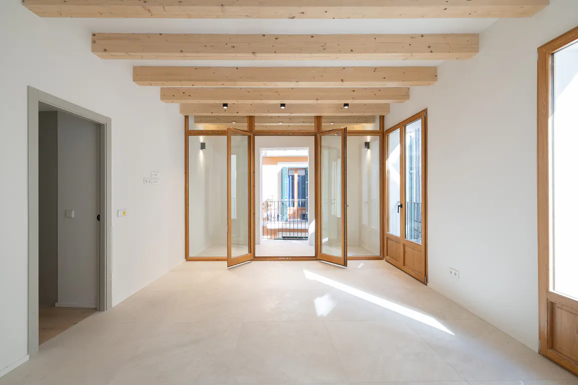 Bright, empty room with white walls, light wood beams, and folding glass doors leading to a balcony. A gray door frame is on the left.