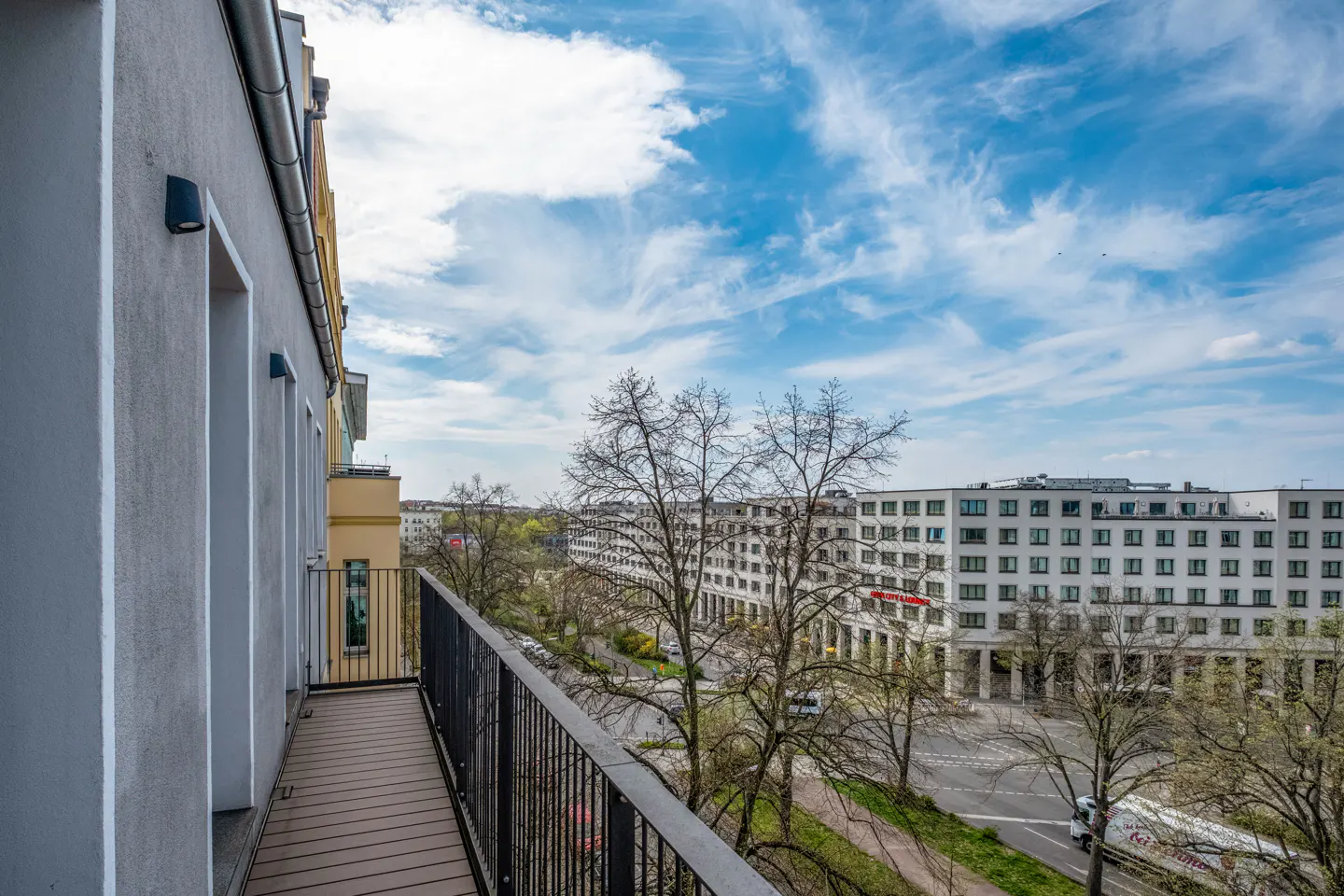 View from a balcony with a black railing, overlooking a city street with white buildings and a blue sky.