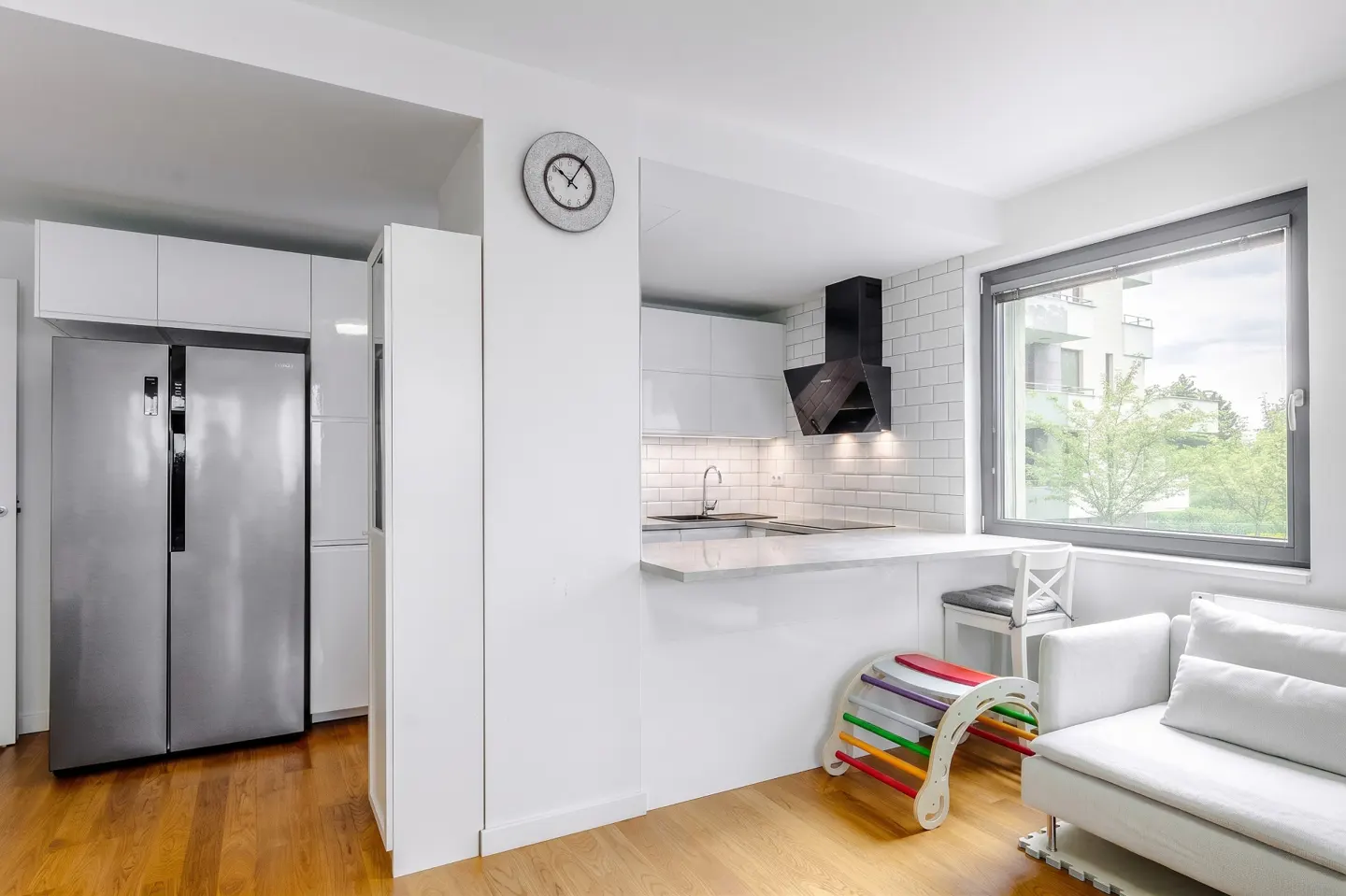 Modern white kitchen with stainless steel fridge, breakfast bar, and a small white sofa with a colorful wooden toy.