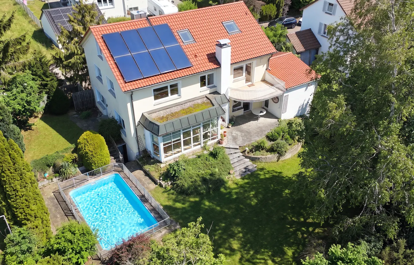 Aerial view of a two-story cream-colored house with a red tile roof and solar panels, surrounded by a green lawn, trees, and a blue swimming pool.
