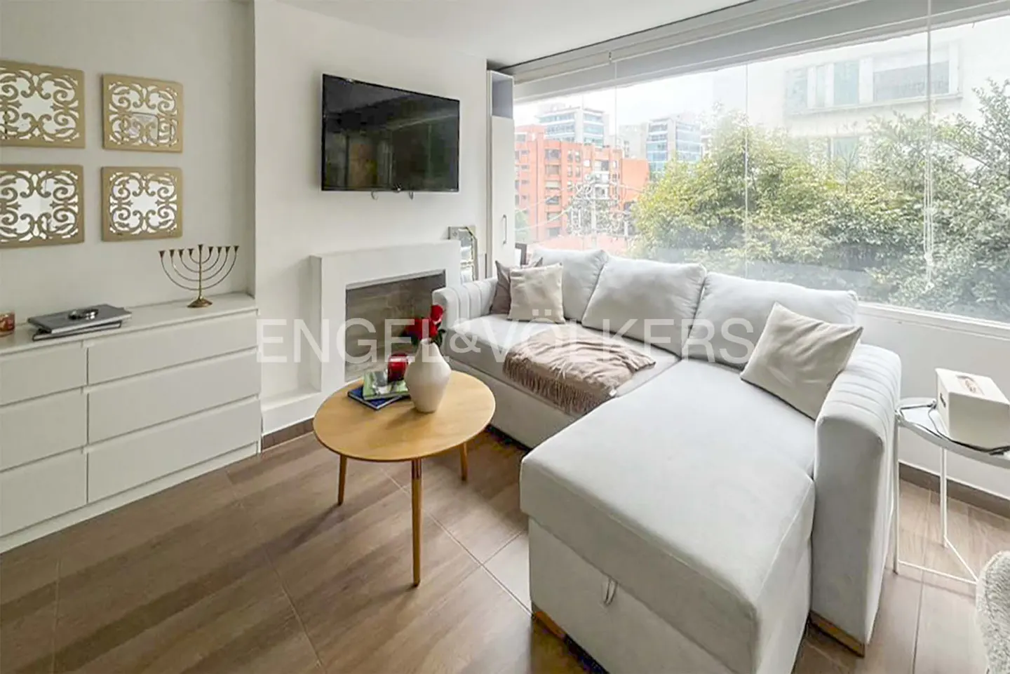 Bright living room with a white sectional sofa, wood floors, and a large window overlooking a city. A round wood table sits in front of the sofa.