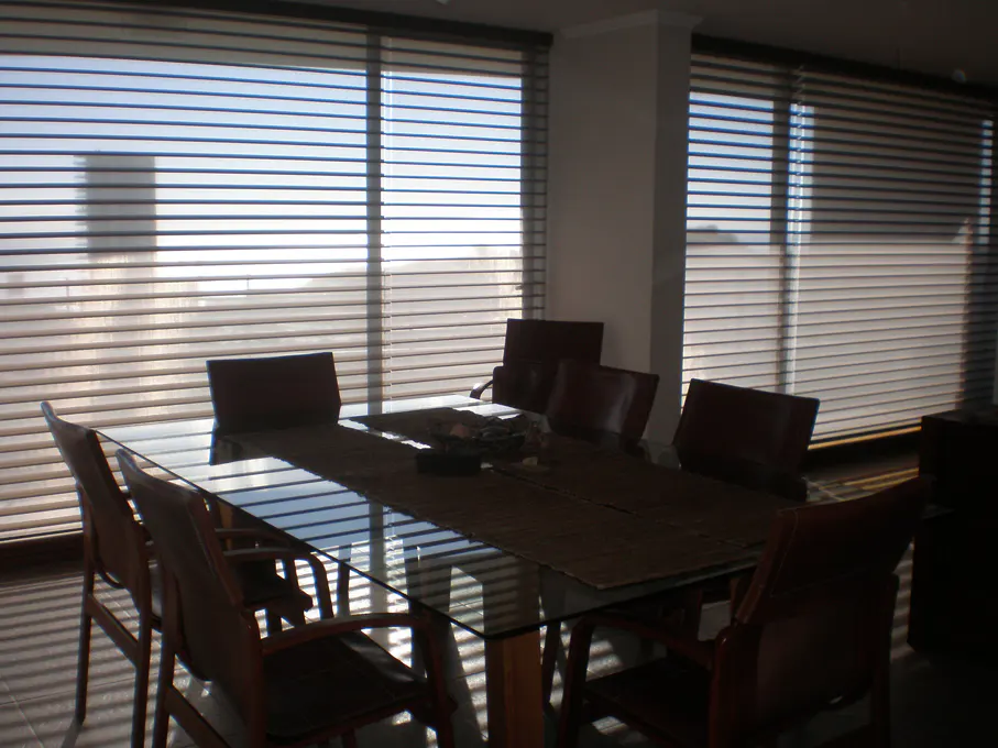 Dining room with a glass table, brown runner, and brown leather chairs. Large windows with horizontal blinds let in natural light.