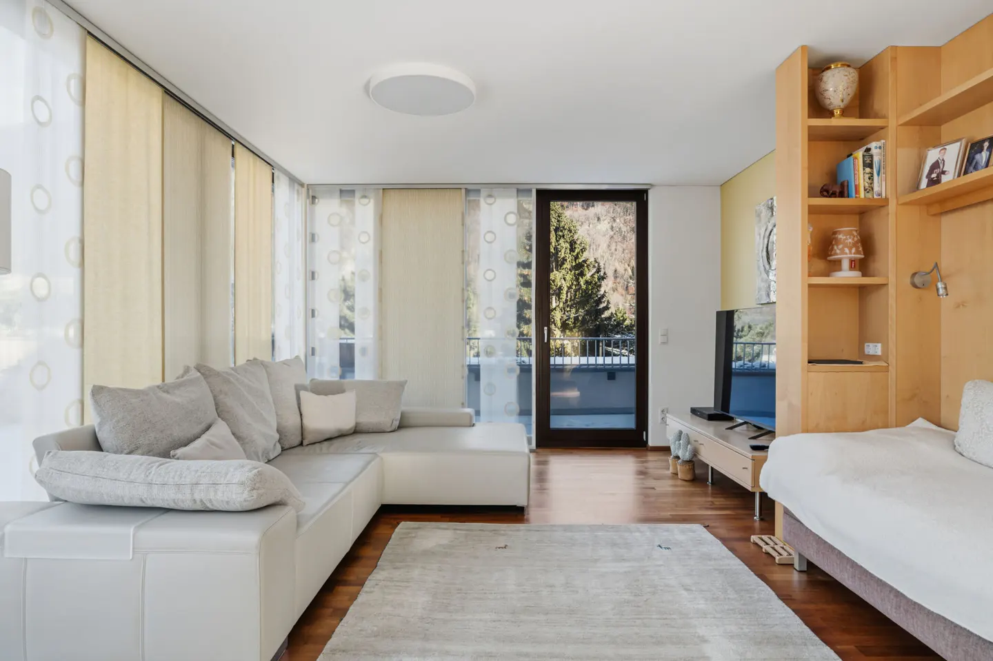 Bright living room with a white sectional sofa, wood floors, and a balcony with mountain views. A built-in bookshelf and bed are on the right.