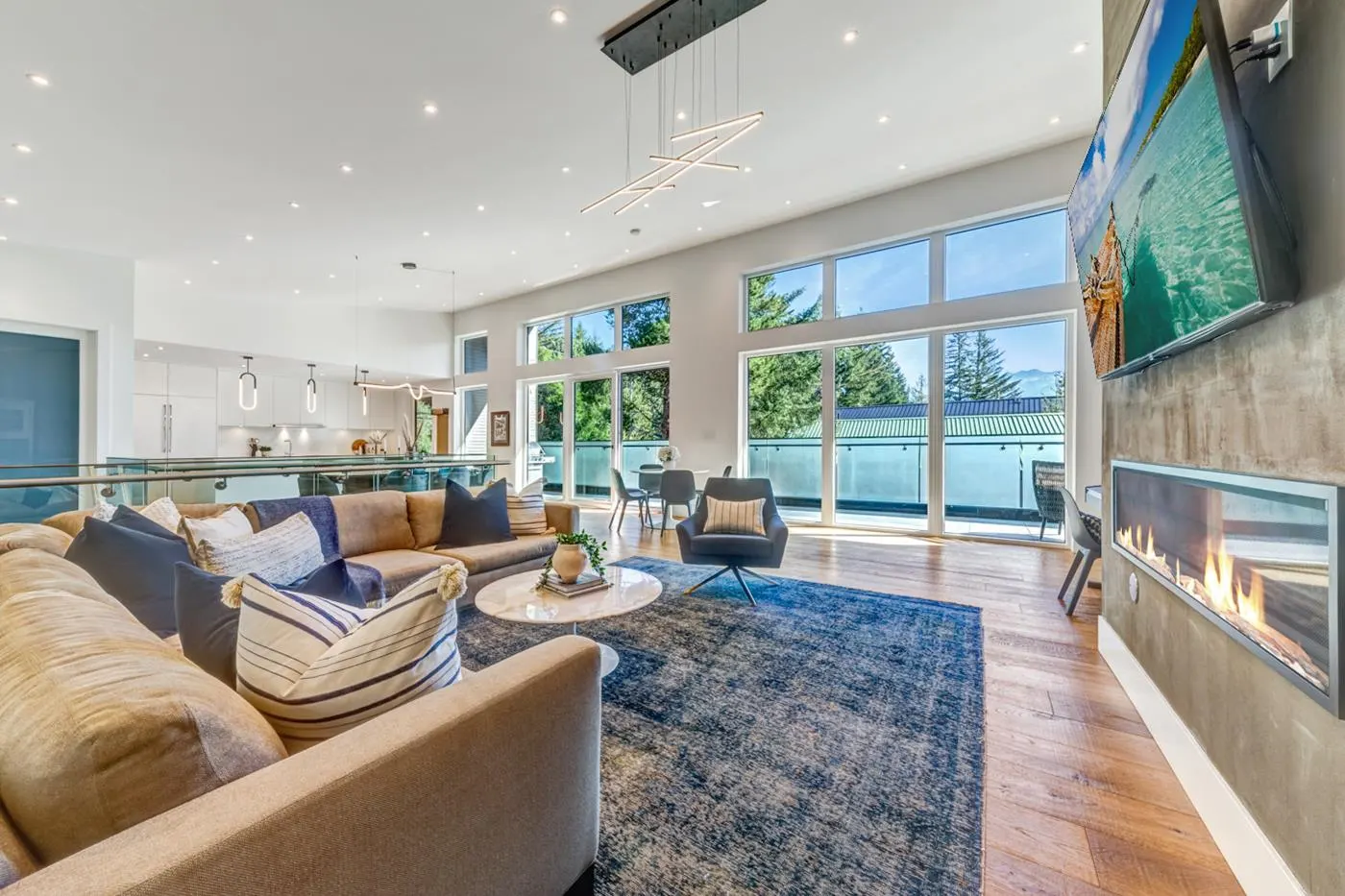 Bright, open living room with a tan sofa, blue rug, fireplace, and large windows overlooking trees. A modern light fixture hangs from the ceiling.