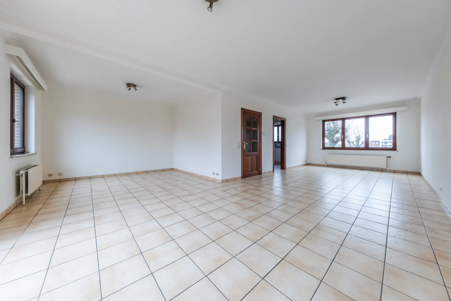 Bright, empty room with white walls, beige tile floor, and brown wood trim around windows and doors.