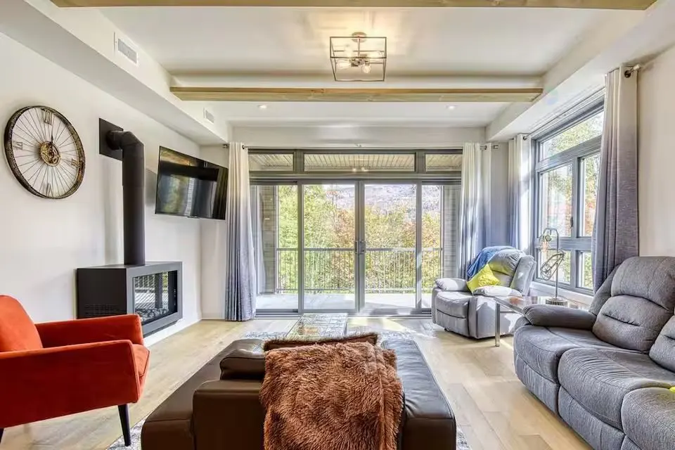 Bright living room with a black fireplace, orange chair, and gray sofa. Large windows overlook trees. A brown ottoman sits in the center.