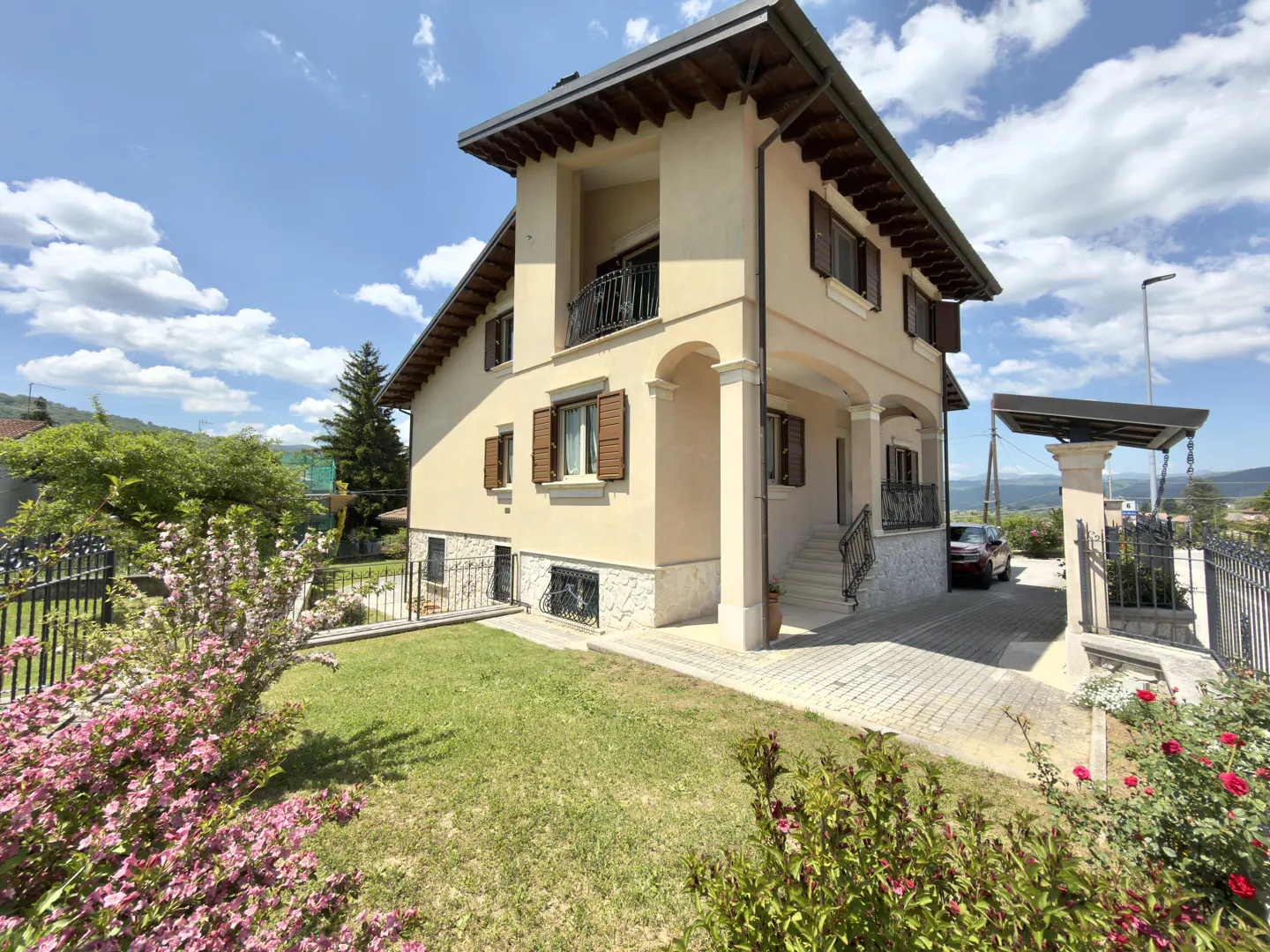 Two-story beige house with brown shutters, stone base, and a paved driveway under a blue sky with scattered clouds.