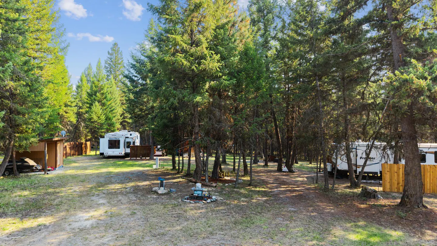 RV park surrounded by tall green trees under a blue sky with white clouds. Two RVs are visible, one on each side of the park.