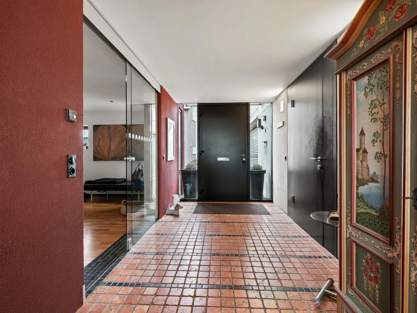 Hallway with terracotta tile floor, red accent wall, and black front door. A painted armoire stands to the right.