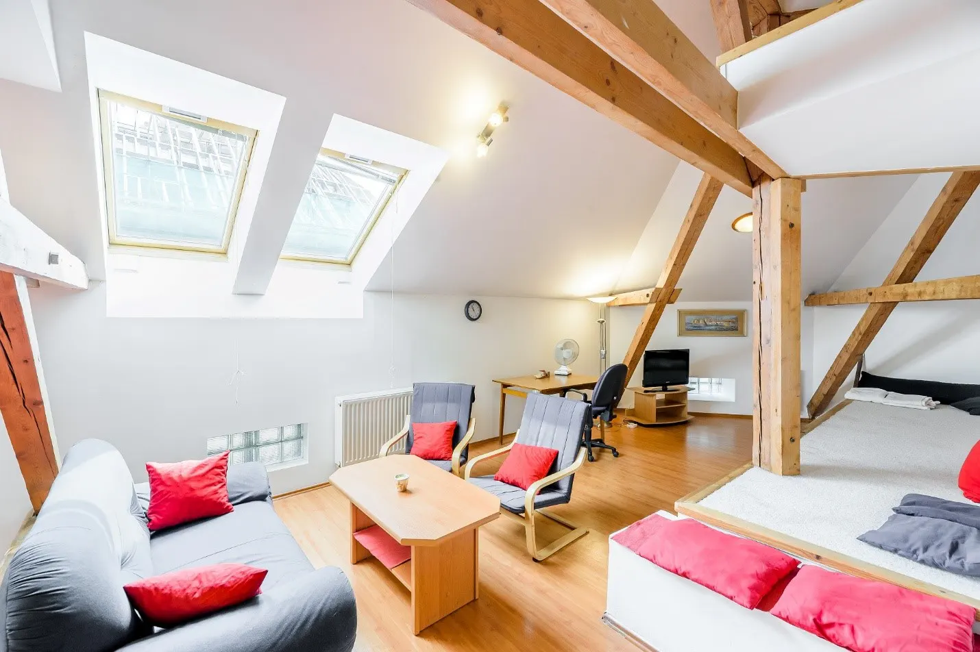 Attic living room with a grey sofa, two armchairs, wooden coffee table, desk, and a raised sleeping area with red pillows.