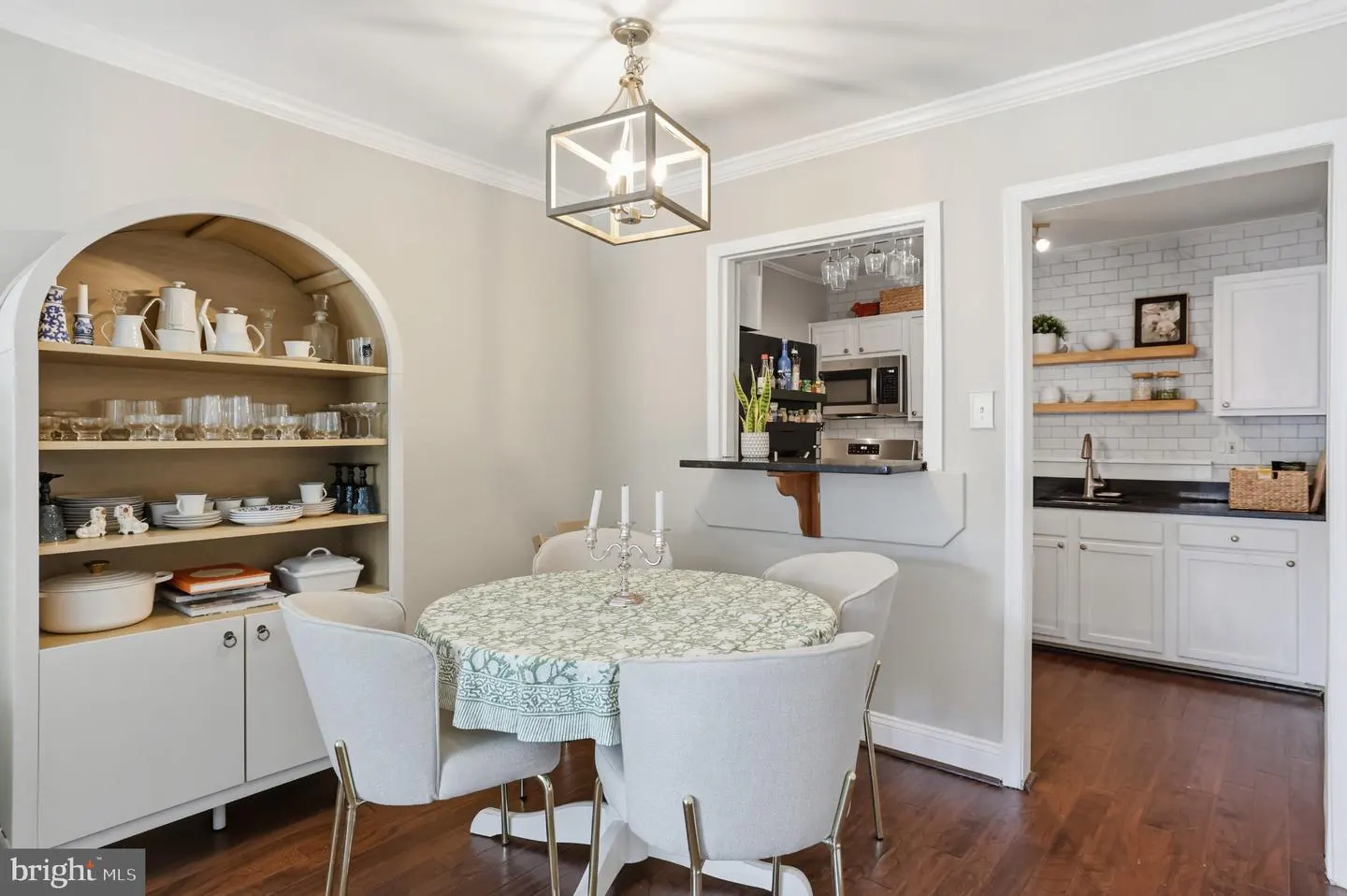 A dining room with a round table, white chairs, a hutch filled with dishes, and a view into the kitchen.