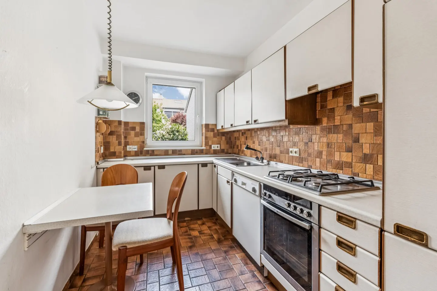 A galley kitchen with white cabinets, brown tiled backsplash, a gas stove, and a small dining area with two wooden chairs.
