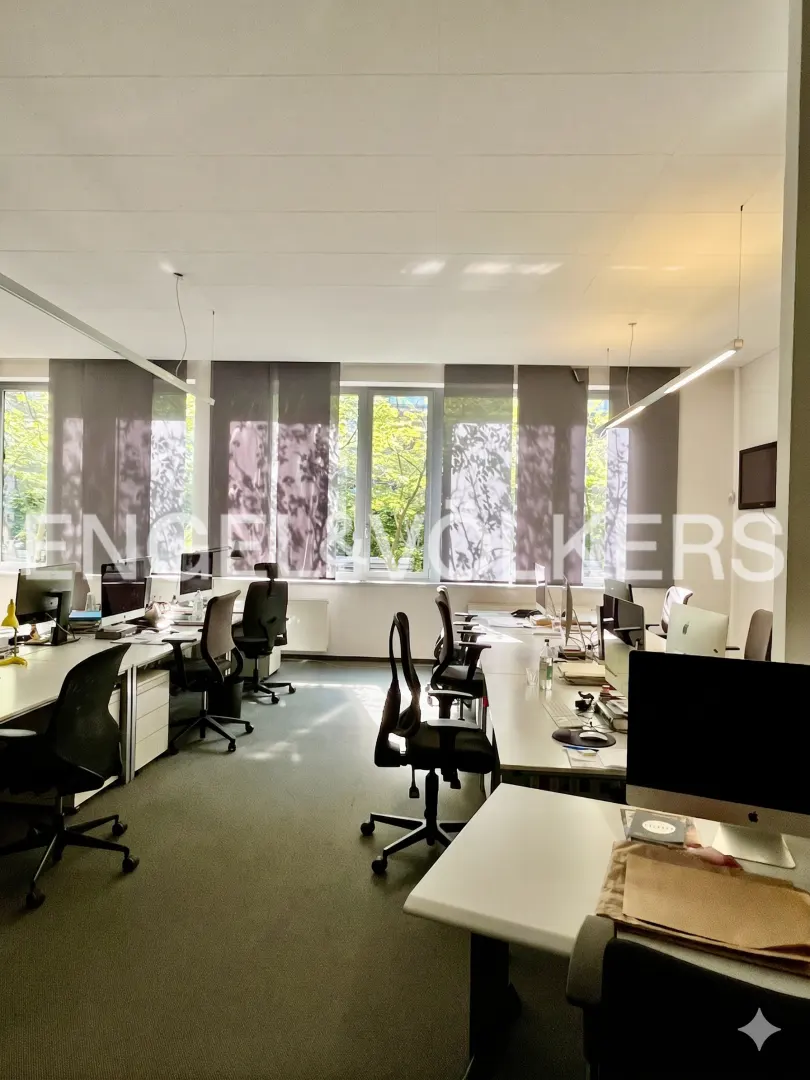 Bright office space with white desks, black chairs, and green carpet. Windows with gray shades overlook trees. "Engel & Volkers" logo visible.