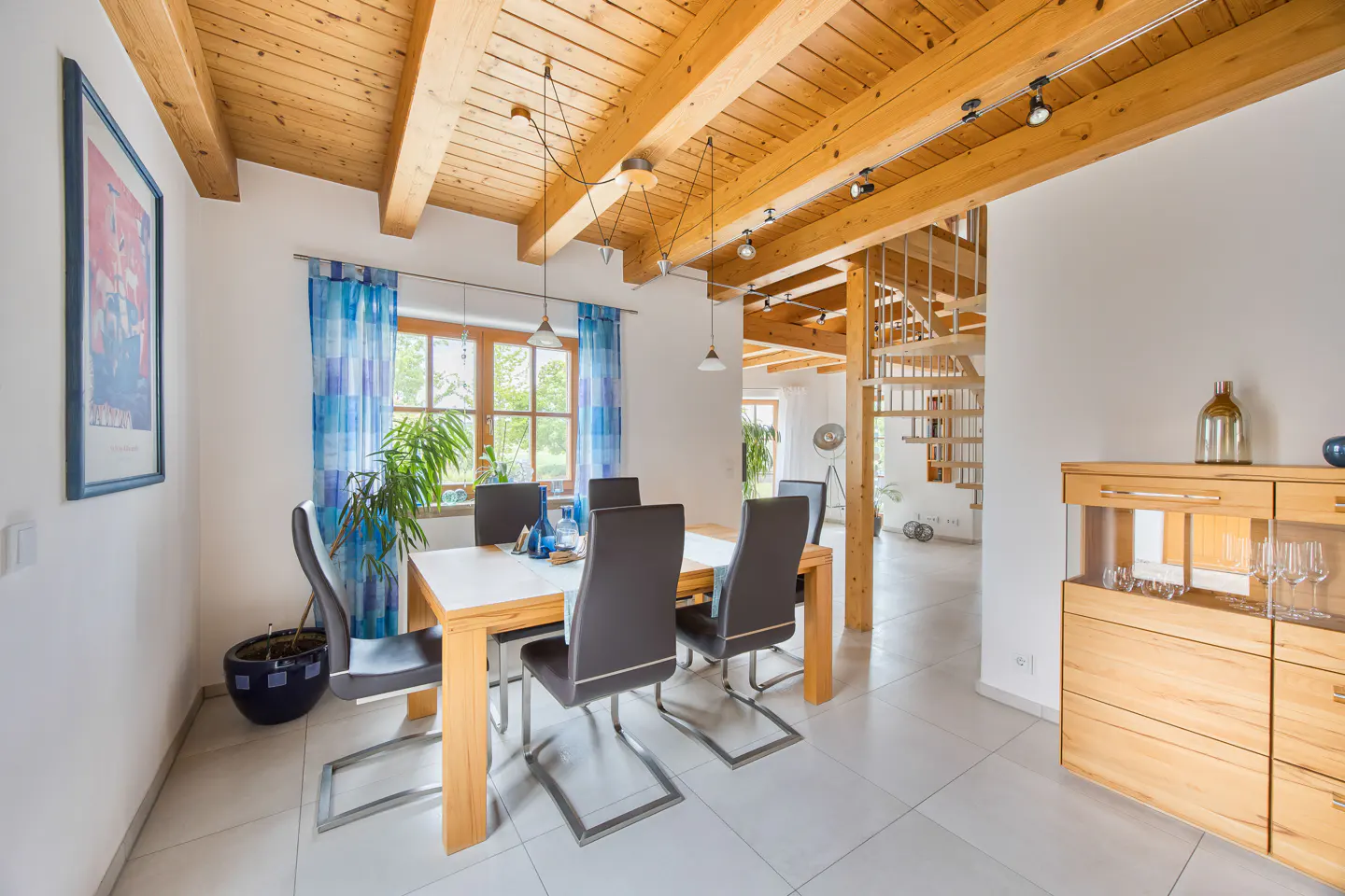 Bright dining room with wood-beamed ceiling, table with six chairs, blue curtains, and a wooden staircase in the background.