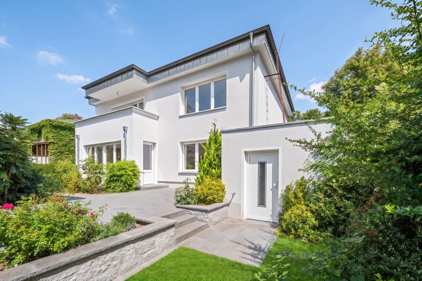 Modern two-story white house with gray roof, stone patio, green lawn, and lush landscaping under a clear blue sky.