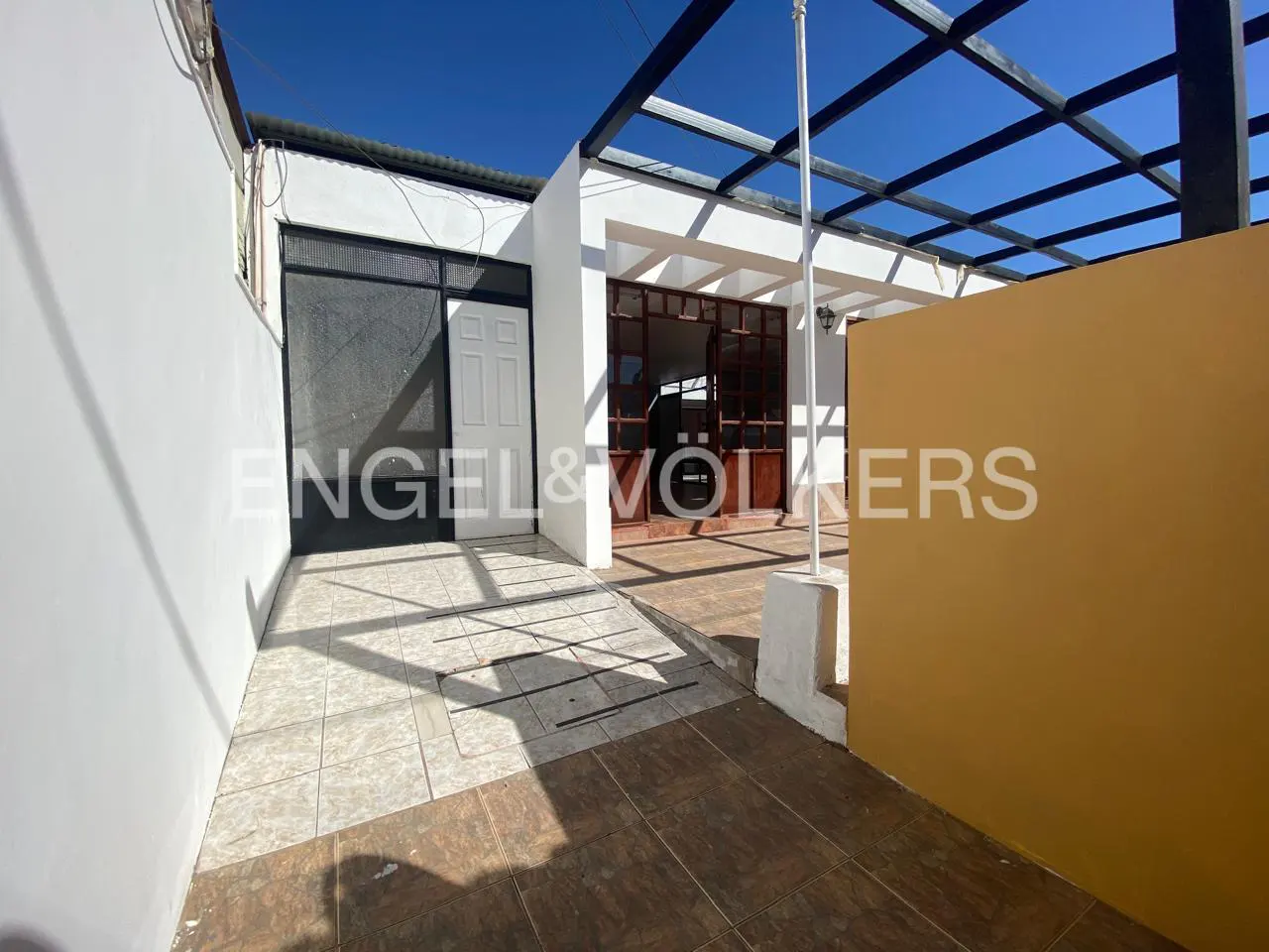 Exterior view of a white house with a black metal pergola and a yellow wall. The ground is tiled. Engel & Völkers logo is visible.