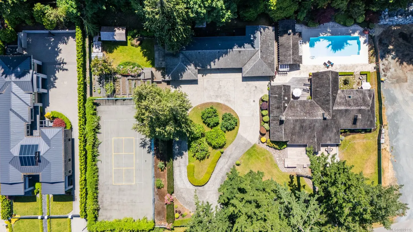 Aerial view of a large property with a house, pool, tennis court, and landscaped grounds. The house has a dark roof and is surrounded by trees.