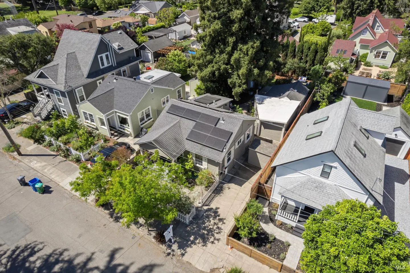 Aerial view of a residential neighborhood with various houses, trees, and a street with recycling bins.