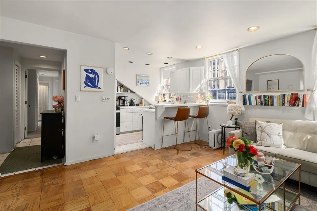Bright living room with parquet floors, white sofa, glass coffee table, and a kitchen breakfast bar with two stools.