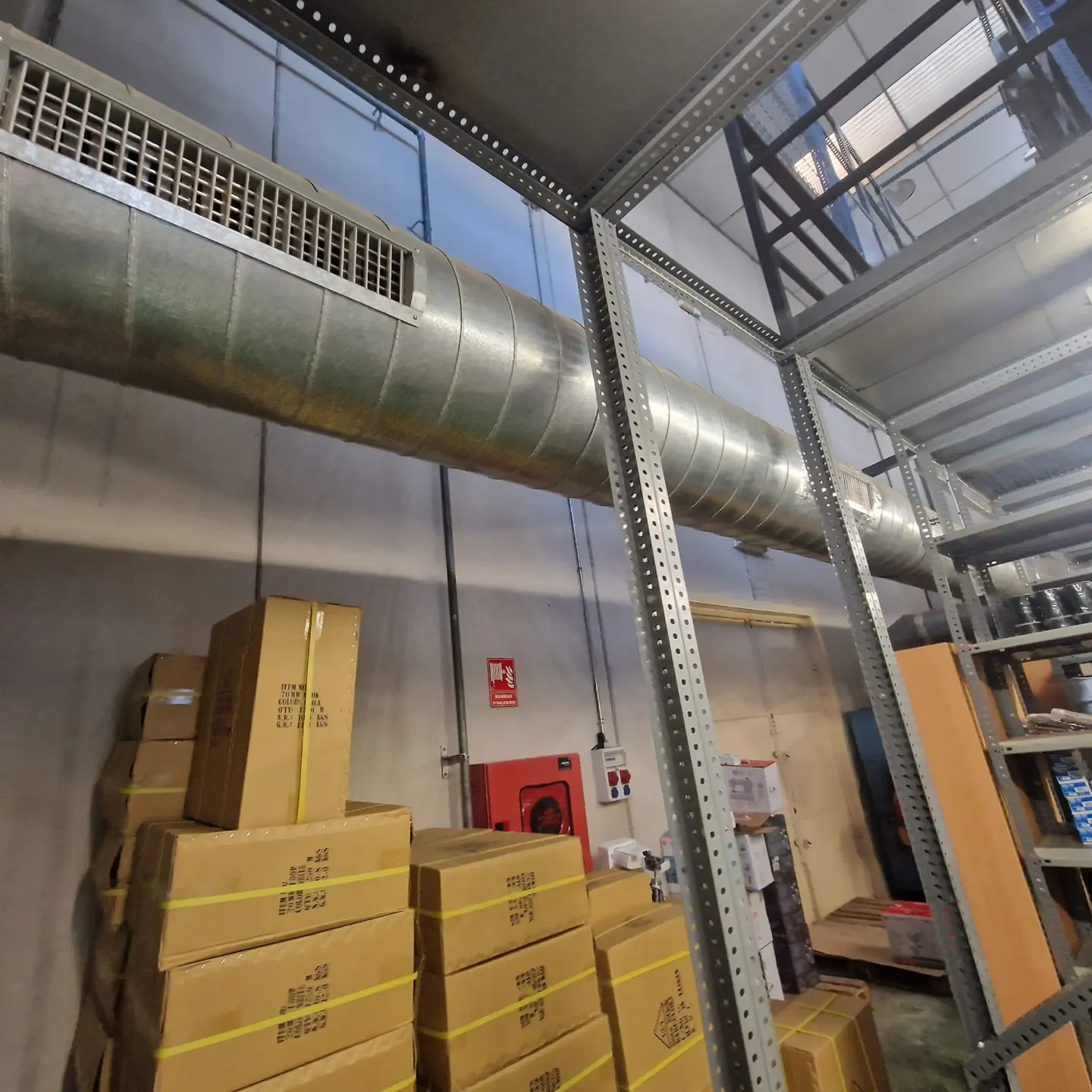 Warehouse interior with stacked cardboard boxes, metal shelving, and a large, silver ventilation duct overhead. A red fire extinguisher is visible on the wall.