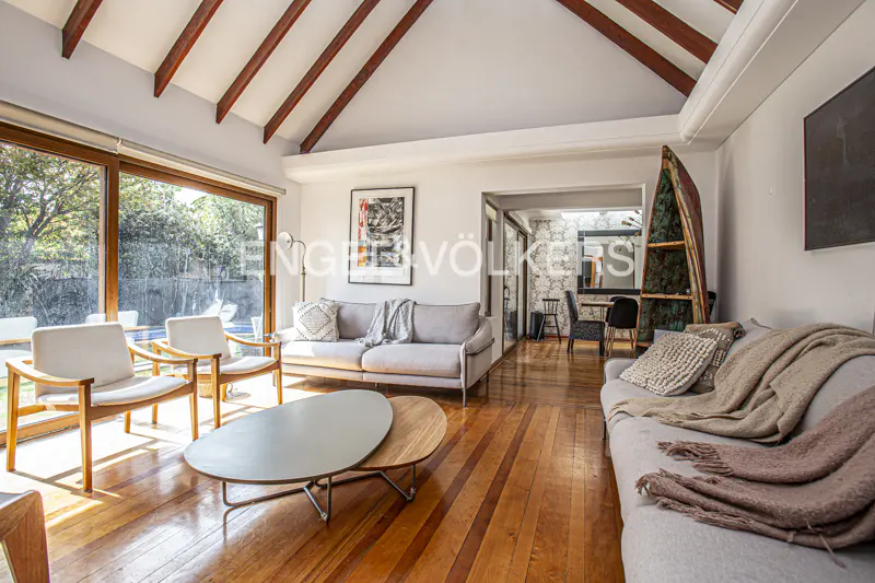 Bright living room with wood floors, vaulted ceiling, and large windows overlooking a pool. Gray sofas and chairs provide seating.