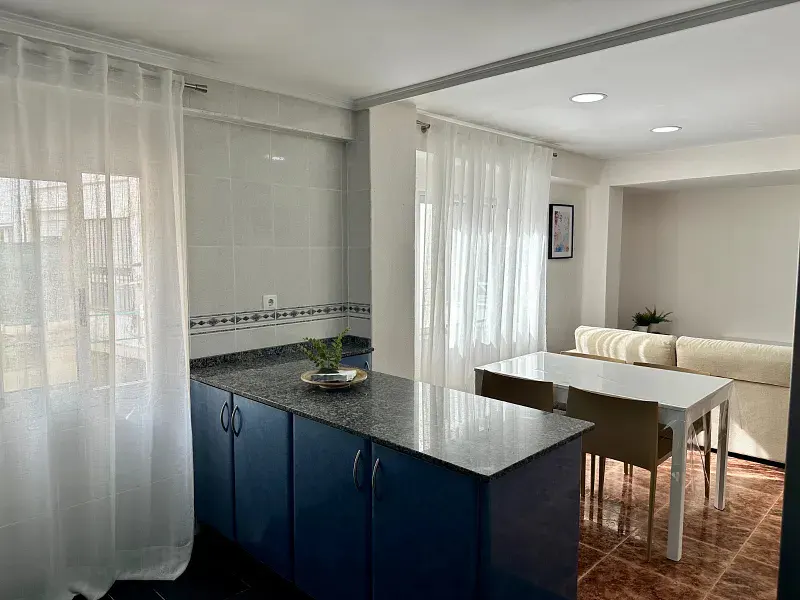 A bright, open-concept kitchen and dining area with blue cabinets, granite countertops, and a white dining table. Sheer white curtains filter sunlight.